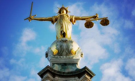 A golden statue of Lady Justice holding a sword in her right hand and scales in her left, mounted on a domed building against a blue sky with clouds.